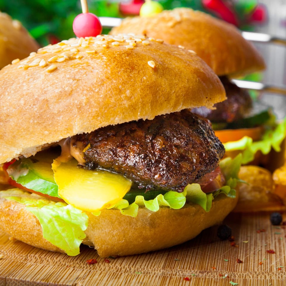Close-up of a mini burger with a sesame seed bun, lettuce, pickles, and a pink skewer.
